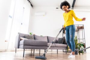 A smiling Black woman with an afro, headphones, yellow sweater, and blue jeans using a vacuum on a wooden floor. The room is bright white and includes a gray, L-shaped couch and a houseplant to the right