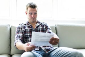 A White man in a flannel shirt sitting on a gray couch who looks surprised as he looks at papers in his hand.