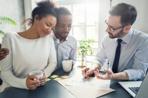 A White man with a beard in a shirt and tie sitting next to a Black man with a beard who has his right arm around a Black woman with her hair in a bun. They're looking at paperwork