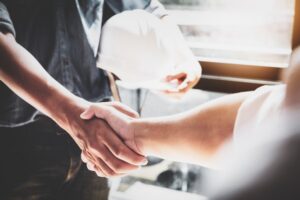 A construction worker shaking hands with another person. The focus is on their hands. The construction worker has a white hard hat in his left hand.