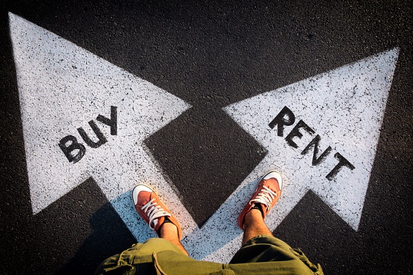 Two white arrows painted on black asphalt. The one on the left says BUY, the one on the right says RENT. There's a person with red shoes and cargo shorts with one foot on each arrow. The view is a point of view of the person looking down at their feet.