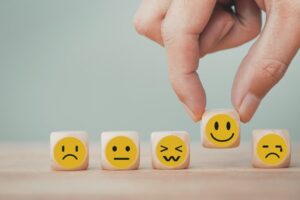 Five wooden blocks in a row, each with a different face on them. A hand is holding the block with a happy face with the thumb and index finger. The block is fourth from the left. The other blocks, from left to right, are "sad face," "indifferent face," "yuck face," and "crying face"