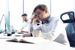 A White man in a white shirt sitting at a computer with a headset around his neck. He's resting his head on his right fist and his cheeks are puffed out. He looks worried.