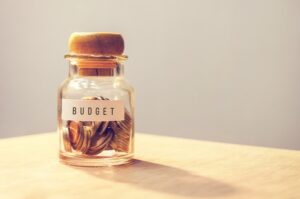 A small glass jar with a cork stopper. Inside the jar are some coins. The word BUDGET appears on a piece f paper taped to the glass. The glass sits on a tan table.