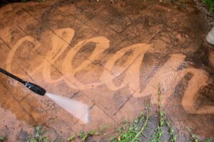 Clay-colored paving tiles being sprayed with a power washer. The power washer is spelling the word "clean" in cursive on the tiles by moving the grime around.