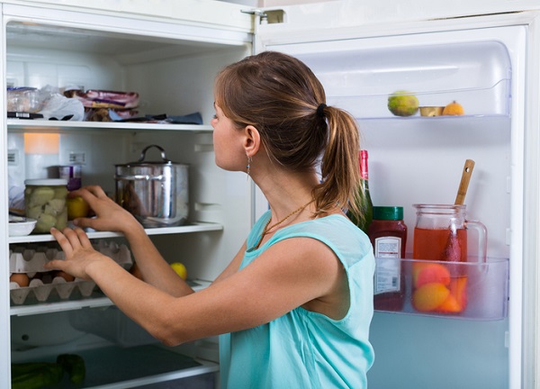 Young woman standing near refrigerator filled with products, thinking about cleaning it